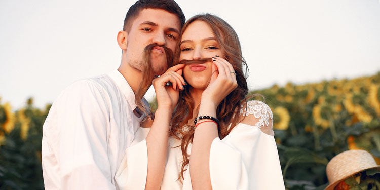 playful couple standing in a field of flowers, both are making mustache from the woman's hair