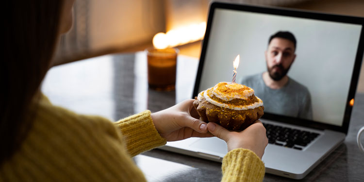 a woman holds a small cake with a candle in front of a computer; the woman is on a video call with her man