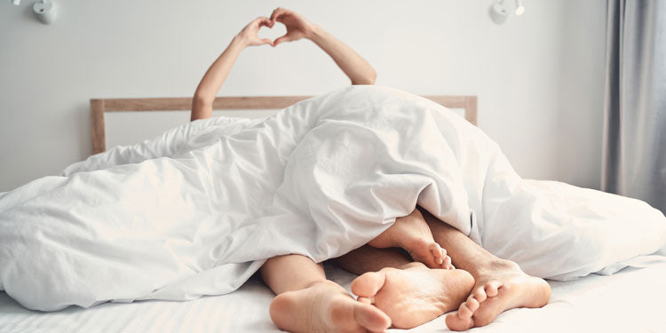 man and woman laying under the sheets; both are holding hands over the bed and forming a heart shape