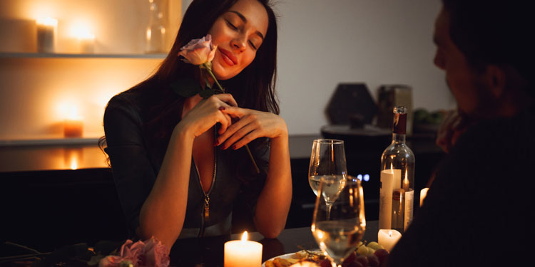 a woman sitting by a table while on a date, she holds a rose and smiles, glasses and candles on the table