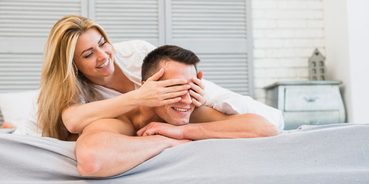 couple laying in bed, the woman is on top of the man's back and playfully closes his eyes