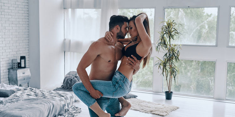 sexy couple posing in the bedroom, both have their shirts off, the man holds the woman's leg by his side