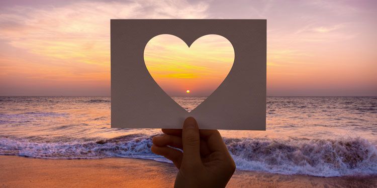 a person holds a heart cutout over a sunset on the beach