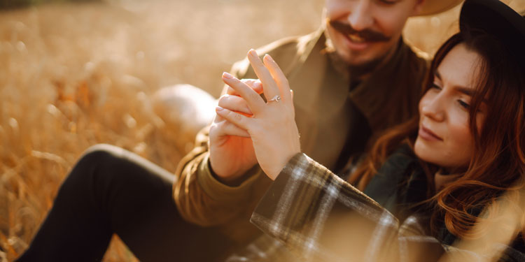 young couple laying down in a field of tall grass, a wedding ring is in the focus