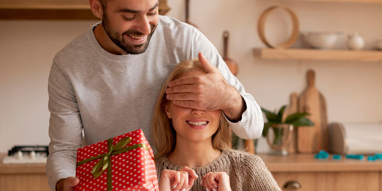 a man surprising his wife with a thoughtful gift, the woman's eyes are closed, both are smiling