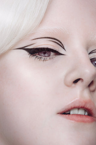 Camryn, an albino Human Beauty model, is show with a close up of half of their face. They are wearing black eyeliner with two lines at the top inner corner of the eye and a short inner corner wing on the eye