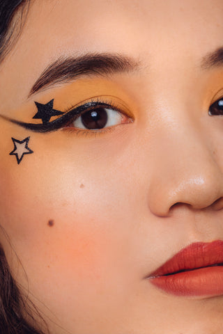 A close up, half face, image of Jen a woman, wearing a yellow base of eyeshadow on her lids with a liner of eyeliner and two black stars near the outer corner of her eye. One is below the liner and is an outliner of a star, the other is a filled in star above the liner and below her eyebrow