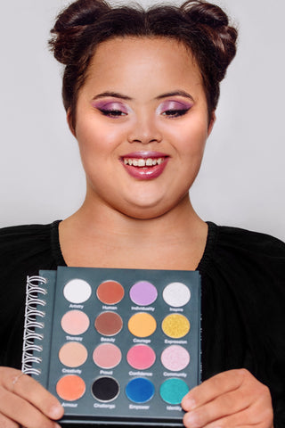 Lauren, a human beauty model, wearing a pink metallic eye look and smiling at the camera. She is holding Human Beauty's Makeup Therapy Palette, open to all the shades, up to the camera