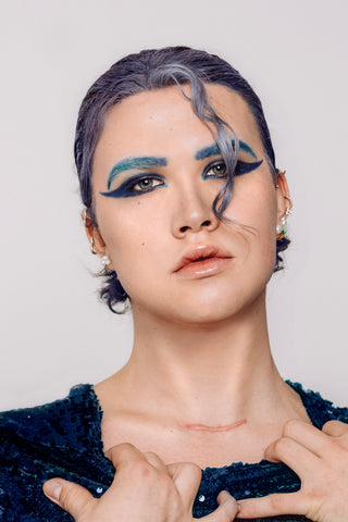 A person with blue hair and eyebrows posing directly into the camera that has bold blue winger eyeliner