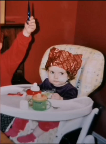 Millie Clare as a young toddler, sitting in a white high chair with two sippy cups infant of her. She has a red Christmas cracker hat on her head and a feeding tube going through her nose.