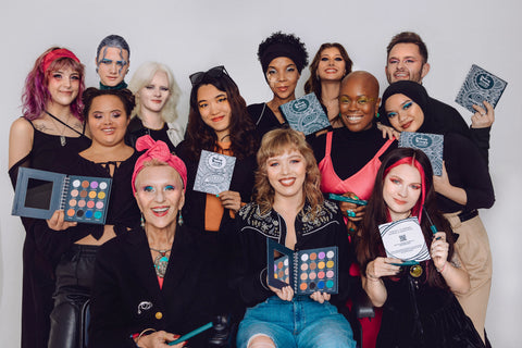 13 people posing with the Human Beauty Makeup Therapy Palette. Each person is wearing black and is smiling at the camera