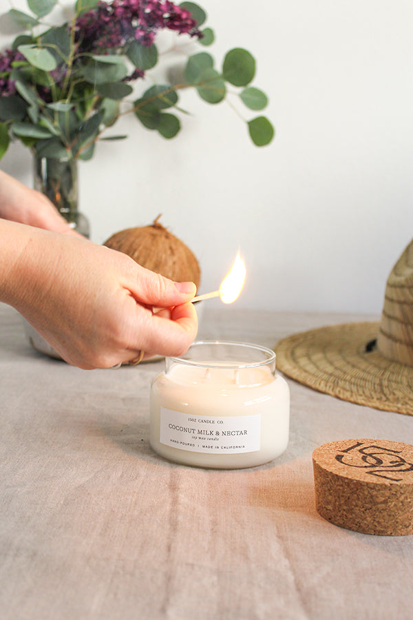 A light skined hand holds a lit match to light a Coconut Milk and Nectar candles with flowers in the background.