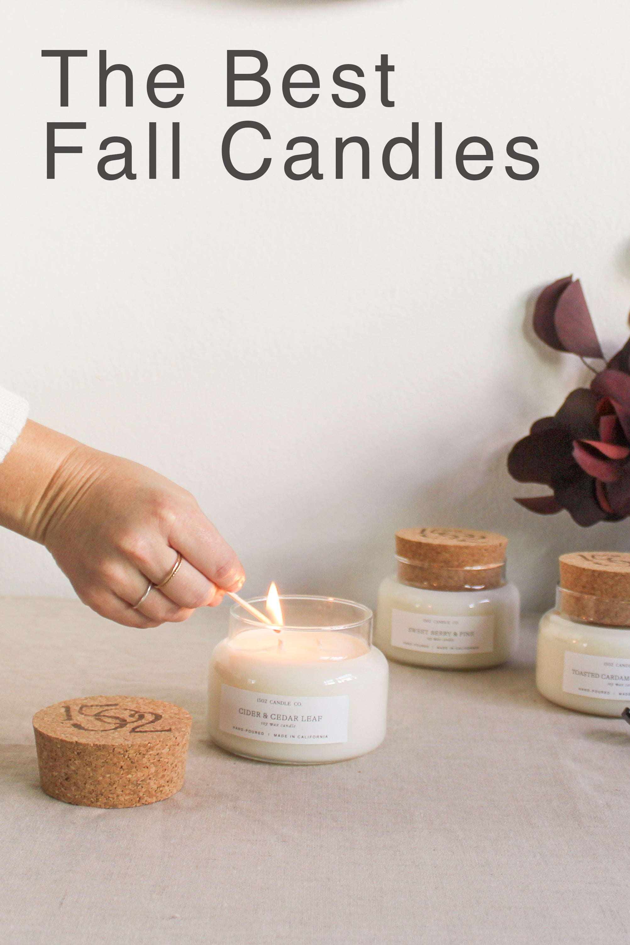 The Best Fall Candles on a minimalist table being lit by a light skined hand with autumn colored folige in the background.