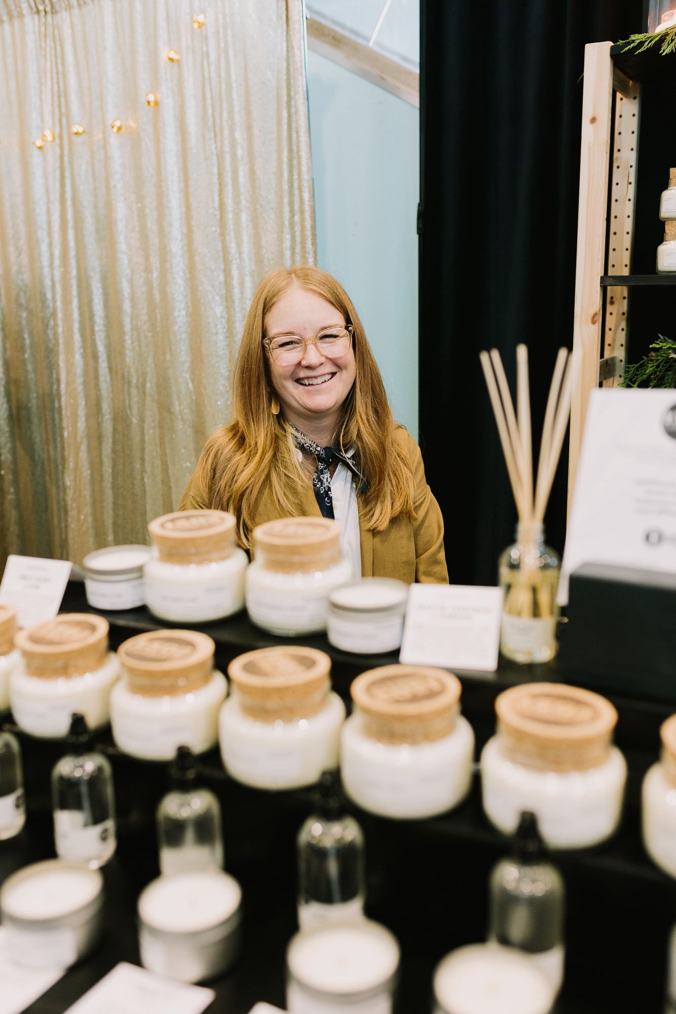 Caroline a fair skined woman with longer red hair and the owner of 1502 Candle Co. smiles behind her candle display.