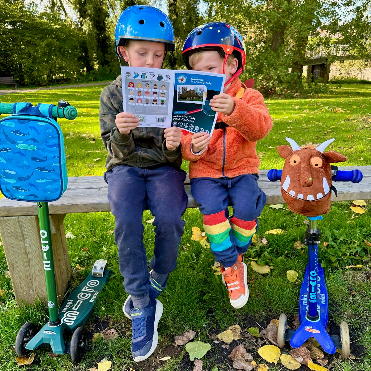 Two children sitting on a bench with their Micro Scooters and Trail booklet