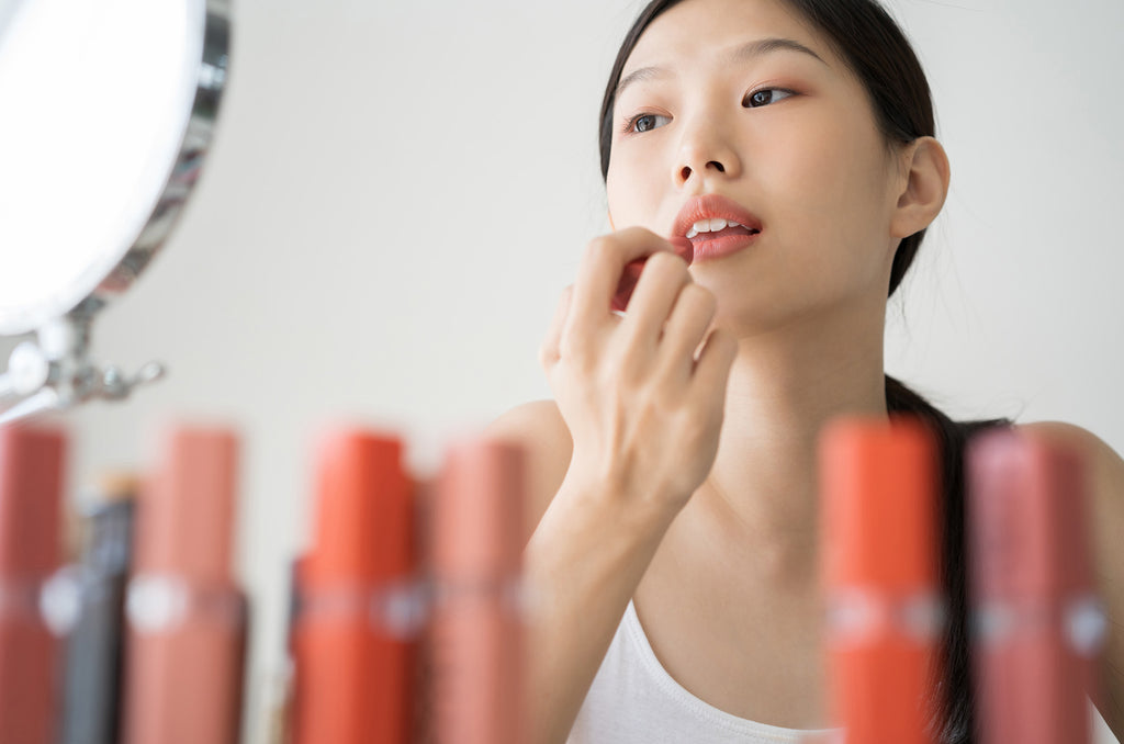 woman putting on lipstick looking at the mirror with lipsticks in the foreground