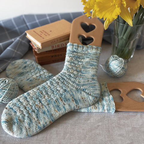 A pair of blue textured hand knit socks lying on a set of old nature books with a vase of bright yellow daffodils. 