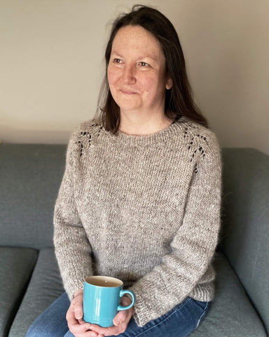 Emma sits on a green sofa with a cup of tea in a blue mug and is wearing a finished Felix sweater that she knit out of Lettlopi yarn