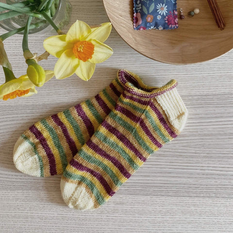 A pair of handknit socks using the Rose City Rollers pattern sits on a table with a vase of daffodils. The socks are striped in yellow, green and purple. 