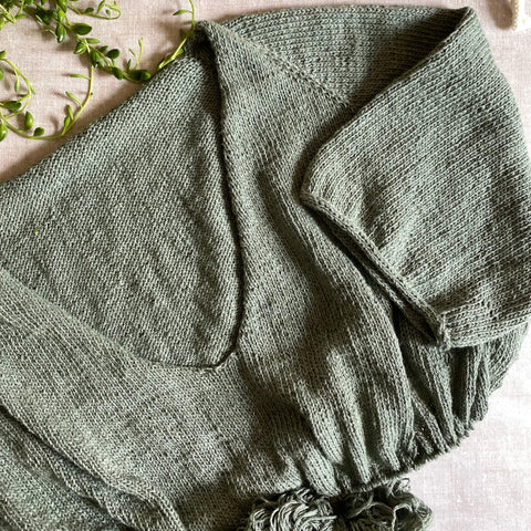 A green handknit t-shirt sits on top of a table covered in a linen cloth. The t-shirt is in mid-knit. 