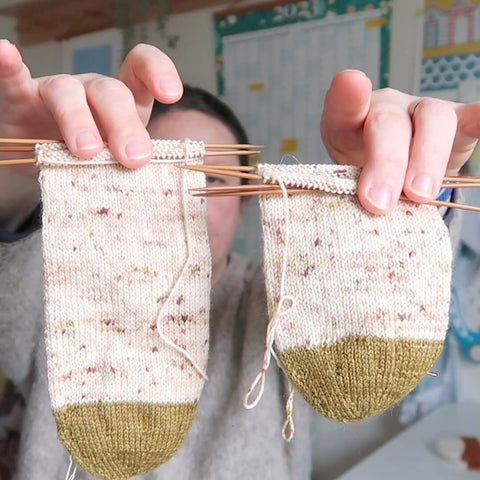 A half knit pair of socks is being shown to the camera during the filming of the Eldenwood Craft knitting podcast. They are knit out of a light pink based yarn and the toes are knit using a sludgy green coloured yarn. 
