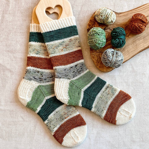 A pair of striped handknit socks in greens, neutrals and a pop of rust, sit on a table covered in a linen cloth. The remnants of the yarn mini skeins used to knit the socks sits next to them ready for their next project. 