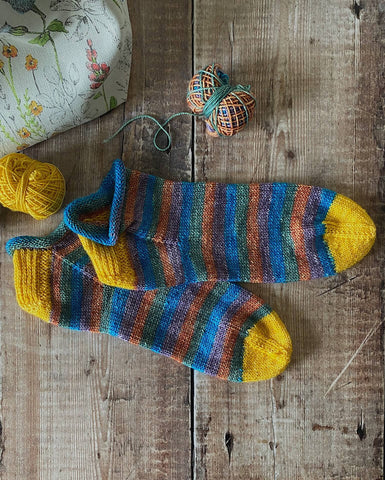 A pair of Rose City Rollers socks hand knit by Emma sits on a wooden table. The socks are striped with bright yellow heels and toes. 
