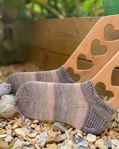 A pair of green, grey and pink striped socks that have been hand knit sit in Emma's vegetable patch on sock blockers. 