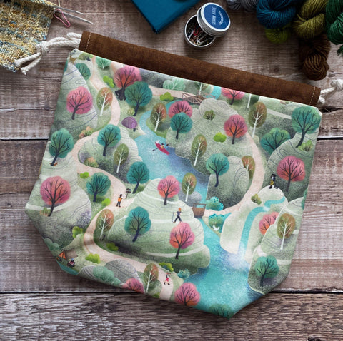 An Everyday Eldenwood Craft project bag lies on a dark wood table surrounded by notions and yarn. The print on the bag shows a scene of hikers walking around hills. 
