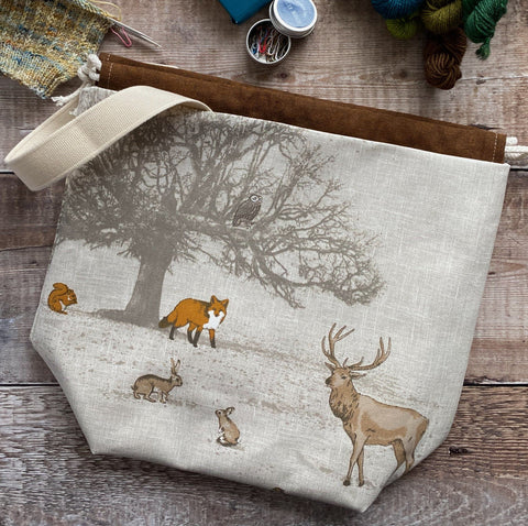 An Eldenwood Craft Midi project bag lies on a dark wood table surrounded by notions and yarn. The scene on the bag fabric shows a winter tree under which can be found foxes, squirrels, rabbits and deer. An owl sits in the tree. 