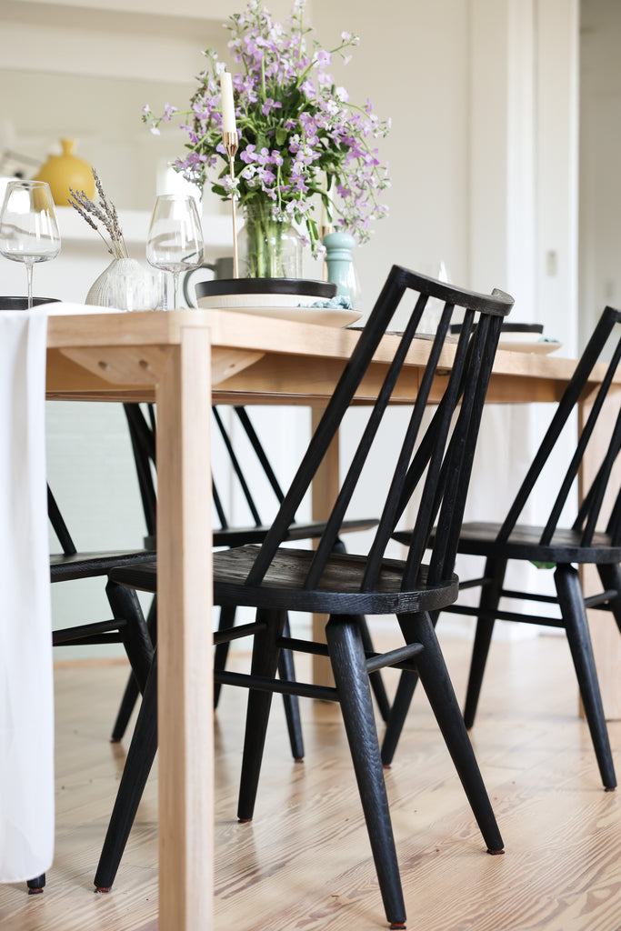 Black farmhouse-style wooden chairs surround a light wood dining table. On the table sits a tall purple flower arrangement, tall tapered candles, wine glasses and place settings.