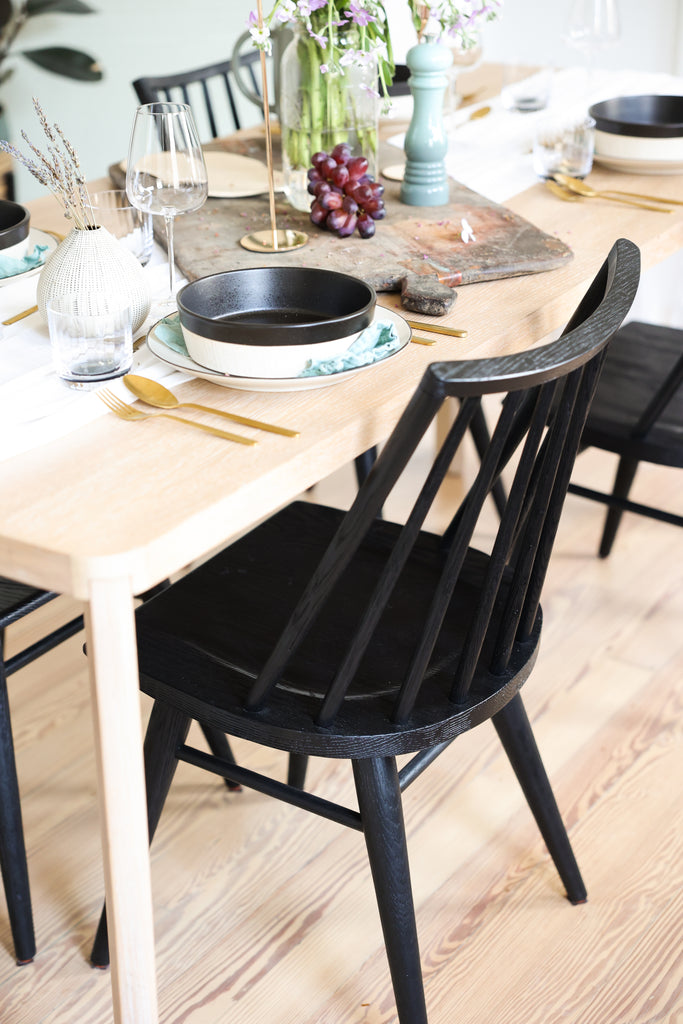 Jenné Claiborne's dining room has a light wood dining table, black farmhouse-style wooden chairs, and black and white dinnerware with gold flatware.