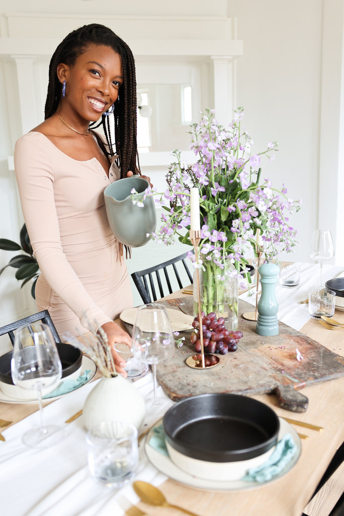 chef and author Jenné Claiborne holds a light teal ceramic pitcher and a clear glass in her dining room, which has a light wood table. On top is a rustic cutting board holding grapes, a tall gold tapered candle holder and candle, and large purple flower arrangement.