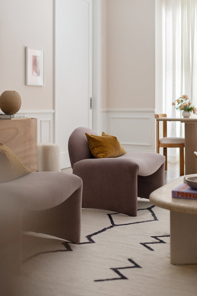 Two velvet armless accent chairs with gold velvet pillows sit on the Moroccan Flatweave rug with a neutral background and black geometric shapes and across from the Marin oval wooden coffee table.