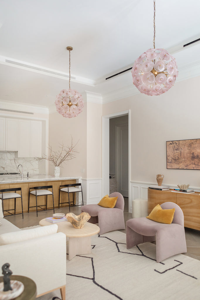 Two pink glass floral pendant lights hang above a modern living room with two armless velvet accent chairs with gold velvet pillows, the oval Marin coffee table, and the Moroccan Flatweave Rug with a neutral background and black geometric shapes.