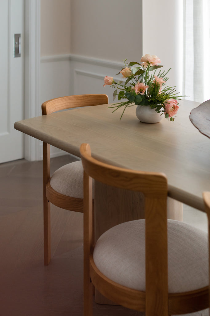 Wooden modern dining chairs with white upholstered seats sit at a light wood rectangular dining table with a small flower arrangement in a white vase.