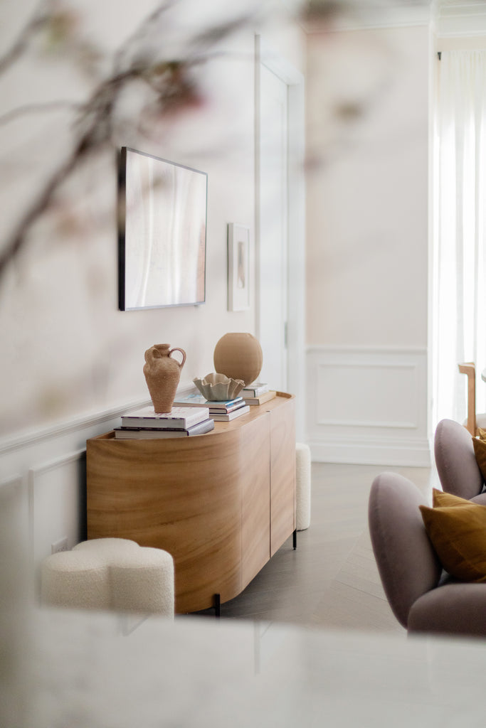 A curved wooden sideboard rests against a wall with stacks of books and three vases. It is flanked on either side with low white boucle clover-shaped ottomans.