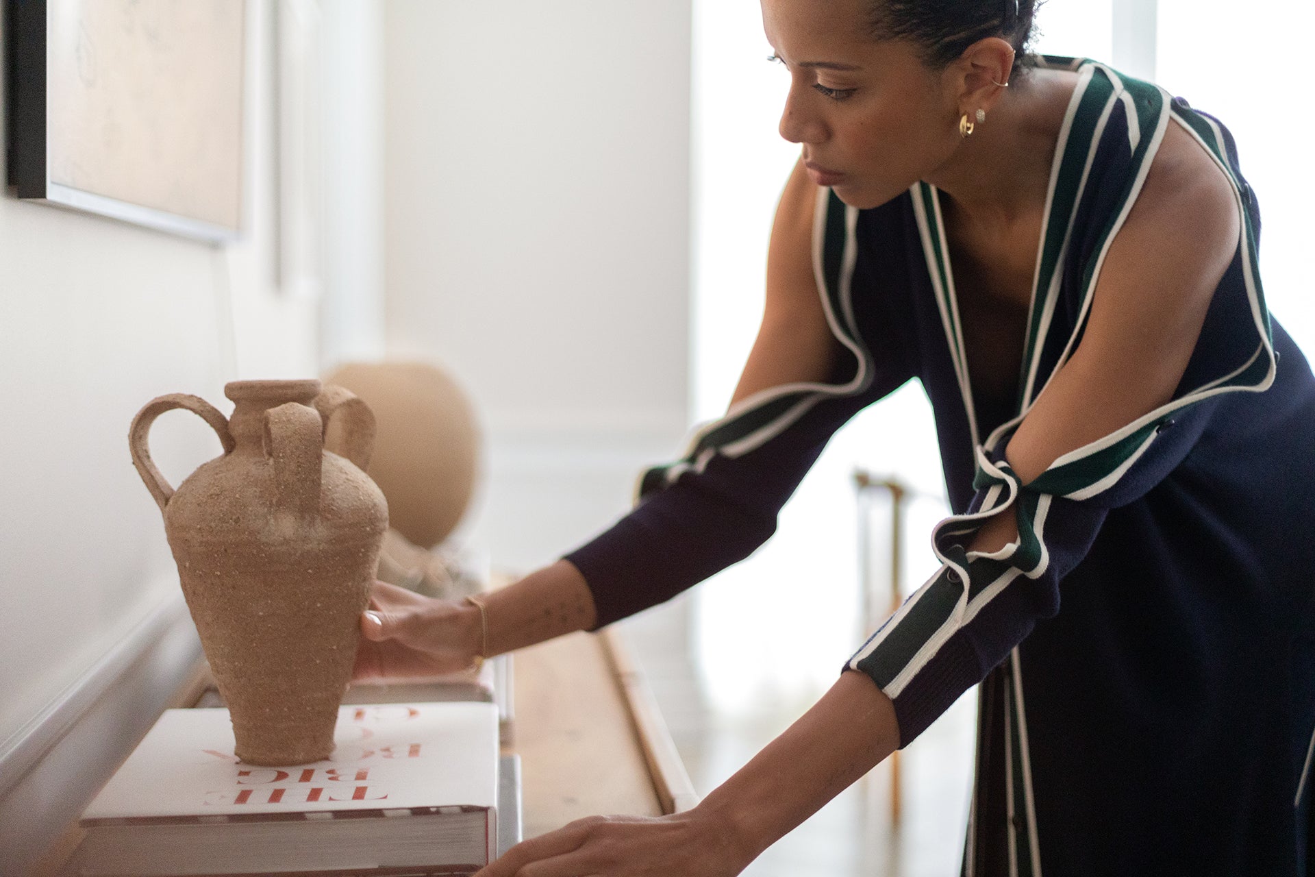 Designer Carly Cushnie - in a navy dress with white accents - adjusts a tall clay vase sitting on books atop a wooden sideboard.