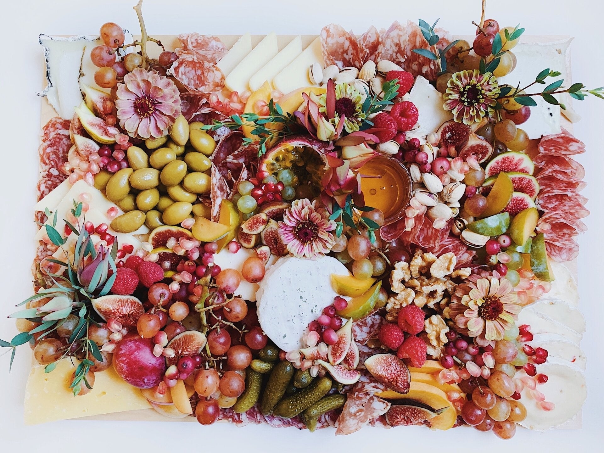 A large charcuterie board features cheeses, meats, olives, fruits, honey and flowers.