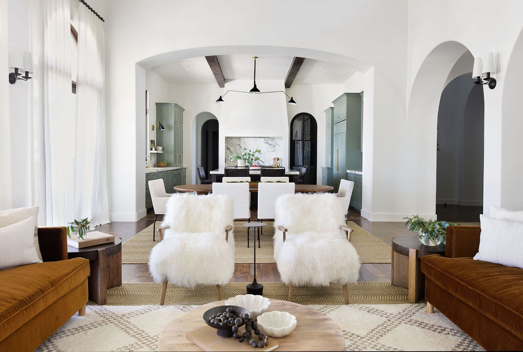 Two fuzzy white Kora arm chairs sit in between two copper Belmont velvet sofas in Jamie Lynn Sigler's home. A chunky light wood round coffee table holds low decorative bowls. A neutral area rug with a brown trellis pattern grounds the space. A dining room table surrounded by white dining chairs and a kitchen are behind the living room.