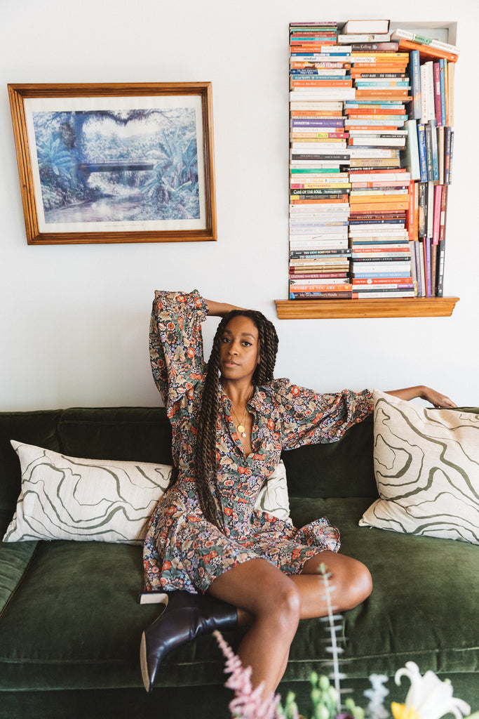 Creator and consultant Chrissy Rutherford lounges on her olive green velvet sofa with ivory and green throw pillows. Above the sofa is a nook holding stacks of books.