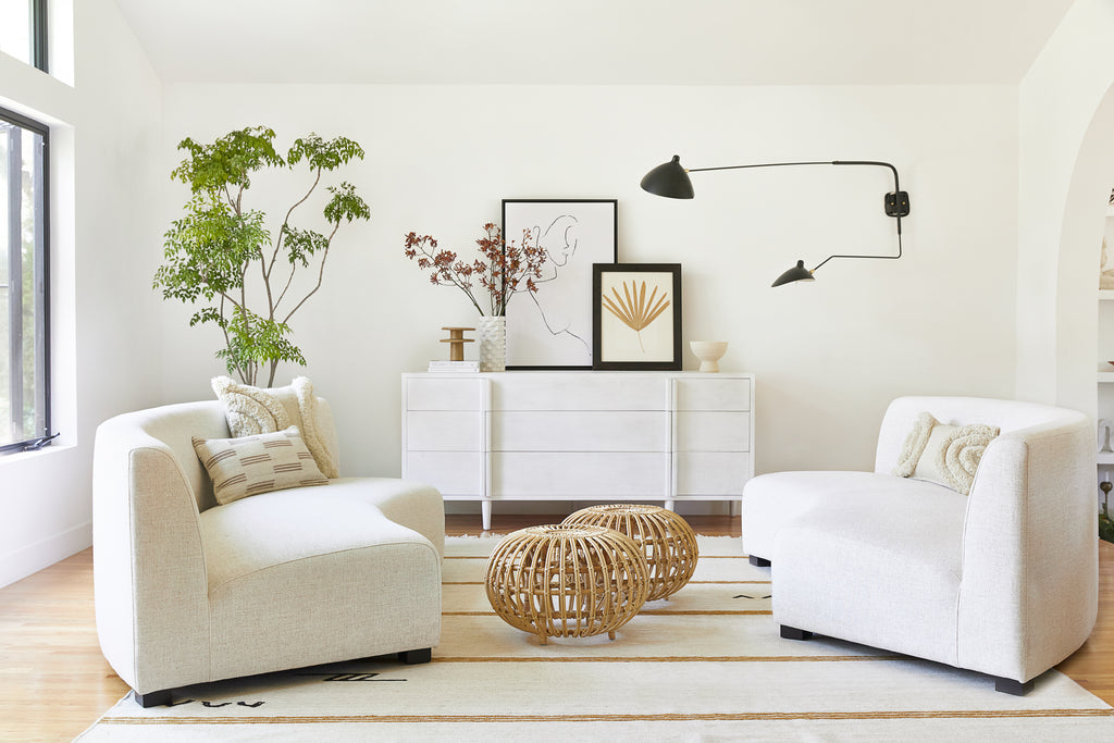 Two rounded ivory linen sofas face each other atop the ivory and gold striped Iconic Stripe Rug by Sarah Sherman Samuel. Two round rattan ottomans sit in between the sofas and a plush fuzzy ivory Arches pillow sits on both sofas while a broken stripe lumbar pillow sits on one.