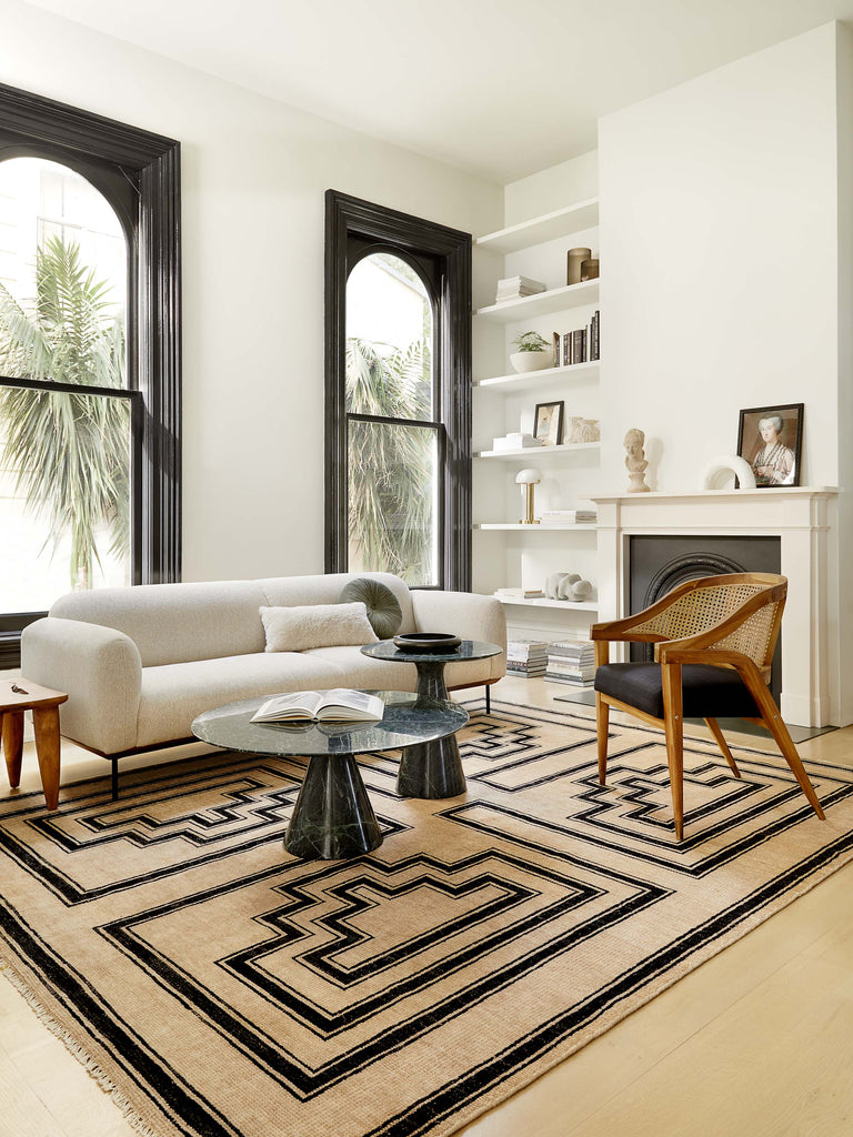 A white modern sofa with curved lines sits atop a neutral and black geometric print Senna area rug. Two round marble tables sit in front of the sofa and a cane and wood rounded arm chair with a black seat sits across from the sofa.
