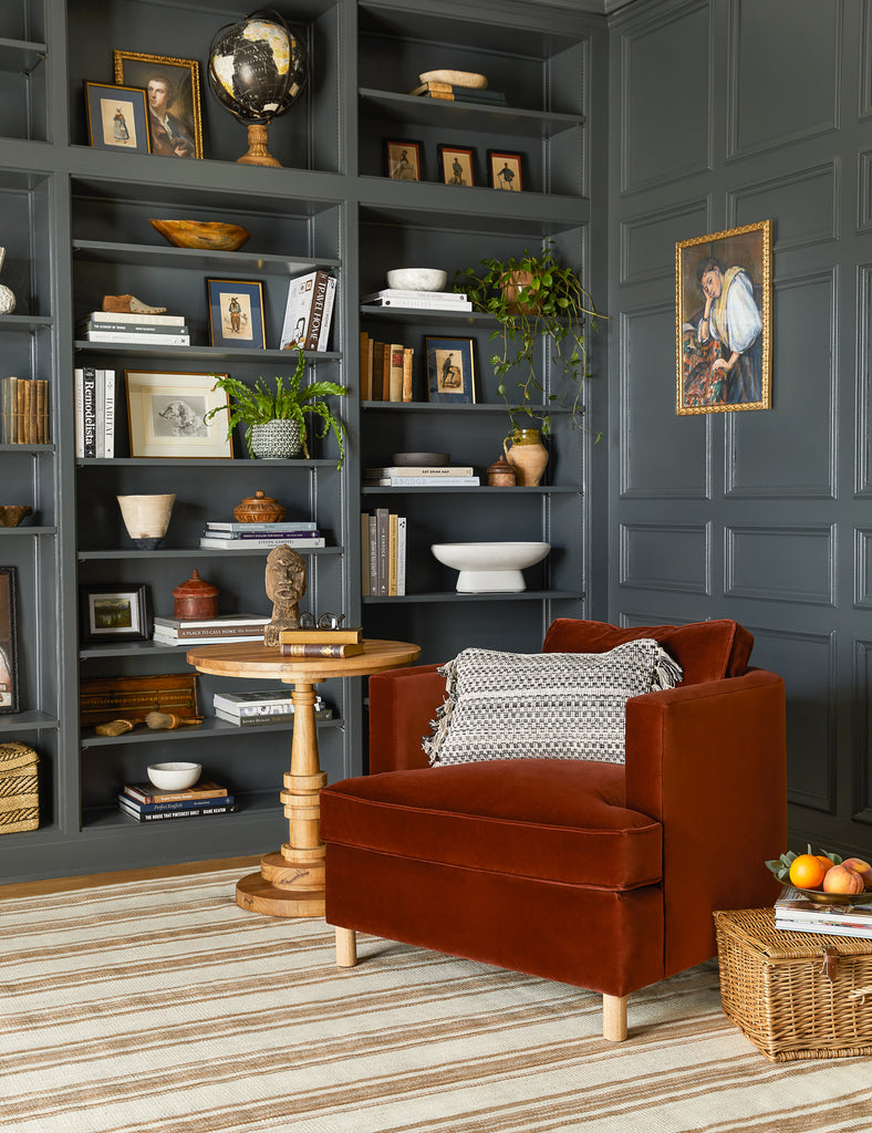 A dark red velvet squared Belmont Accent chair sits in a dark gray paneled library. Next to the chair is a round wood side table and built-in bookcases full of plants, books, and home decor items.