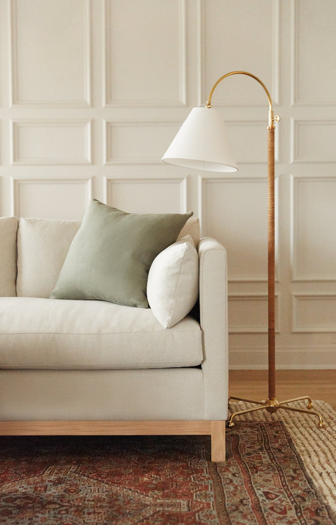 The rattan wrapped Davis gold floor lamp with white linen shade sits next to a light cream sofa with moss green square throw pillow.