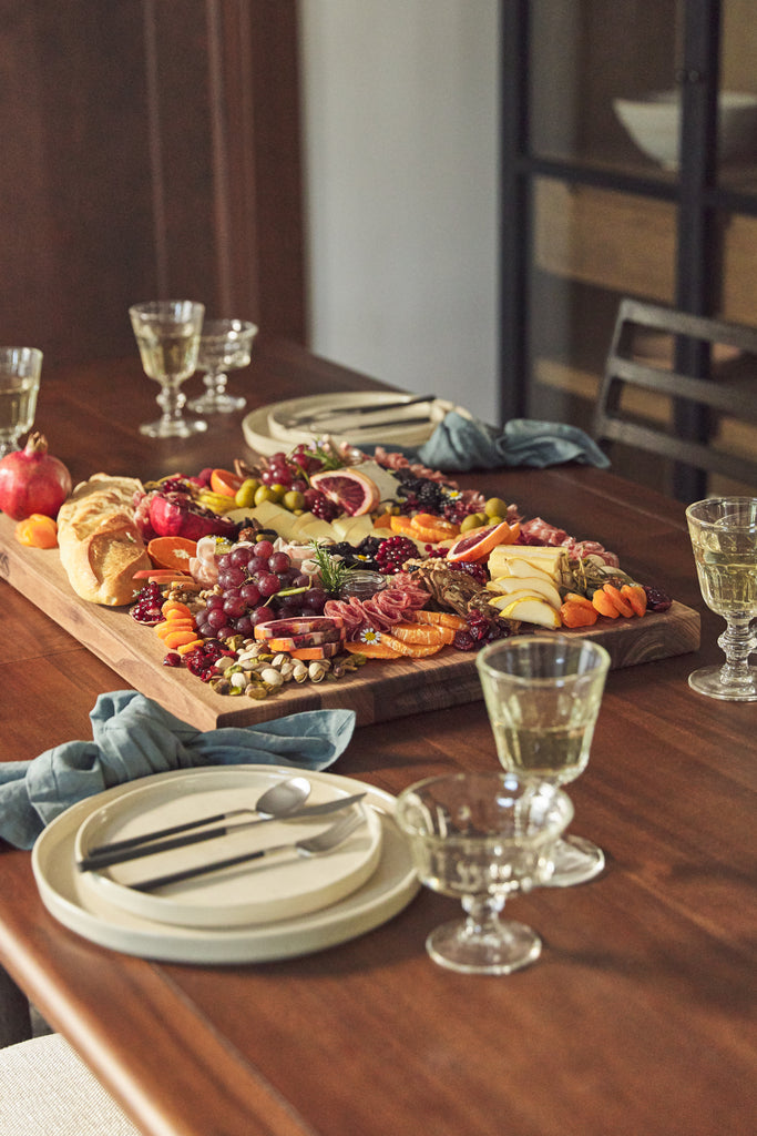 Aimee Lennox's cheeseboard with fruits, cheeses, meats, and nuts is on a wooden board on a wooden dining table surrounded by wine glasses full of white wine.