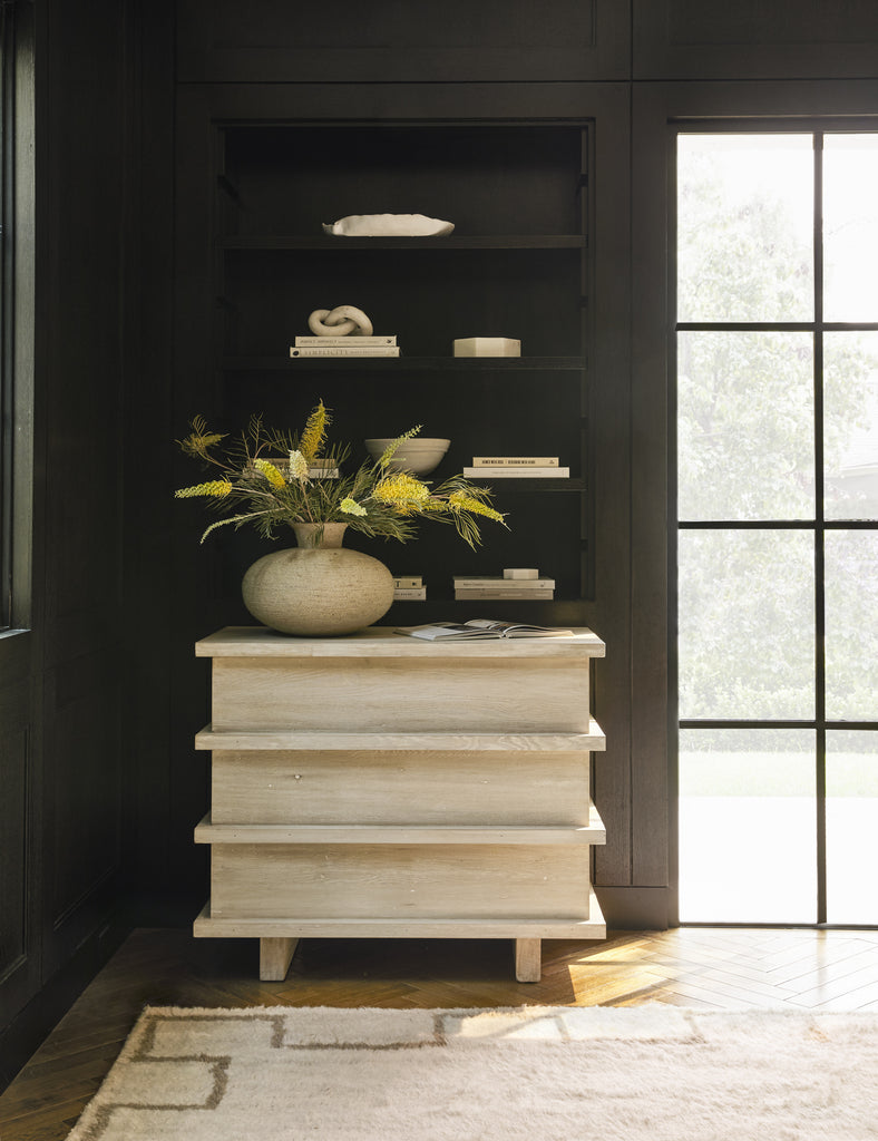 A small light wood dresser with a short round textured vase holding dried botanicals sits underneath a dark hanging bookcase with white books and decorative accents in a modern entryway look.