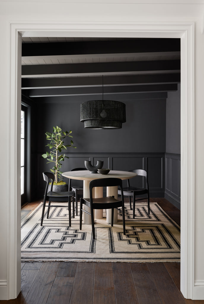 The black woven rattan Sayan pendant light hangs in a black dining room above a round white dining table, rounded black dining chairs and above the neutral and black geometric patterned Senna Rug.