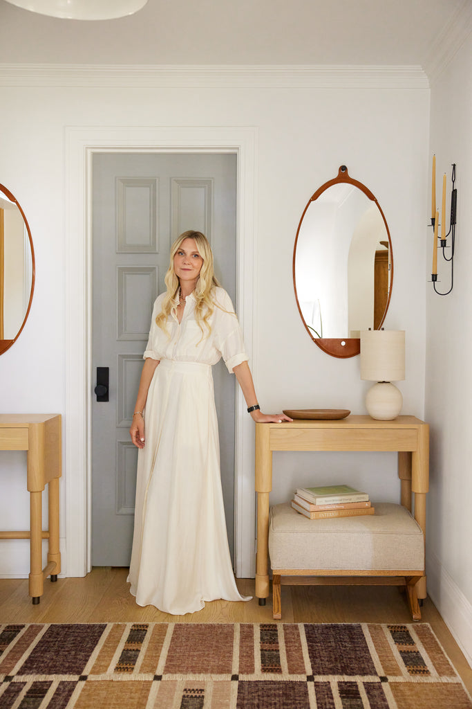 Interior designer Nina Freudenberger stands between two light wood side tables, one with a linen upholstered ottoman underneath. The multicolored neutral woven Anni rug from her exclusive Lulu and Georgia collection lays on the floor.