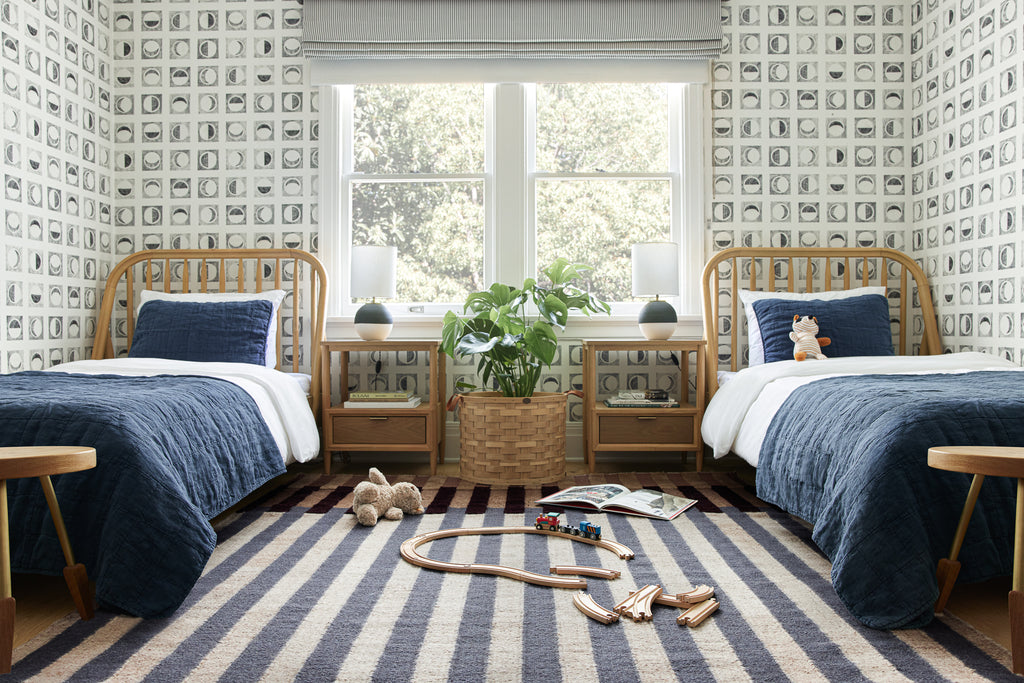 Two twin beds with navy blue bedding and white sheets sit on either side of a window. The neutral woven striped Otti rug lays on the floor and has wooden toys on it.